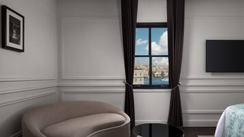 A modern hotel room with a curved sofa, a round table, a large window with curtains, a flat-screen TV, and a framed picture on the wall.
