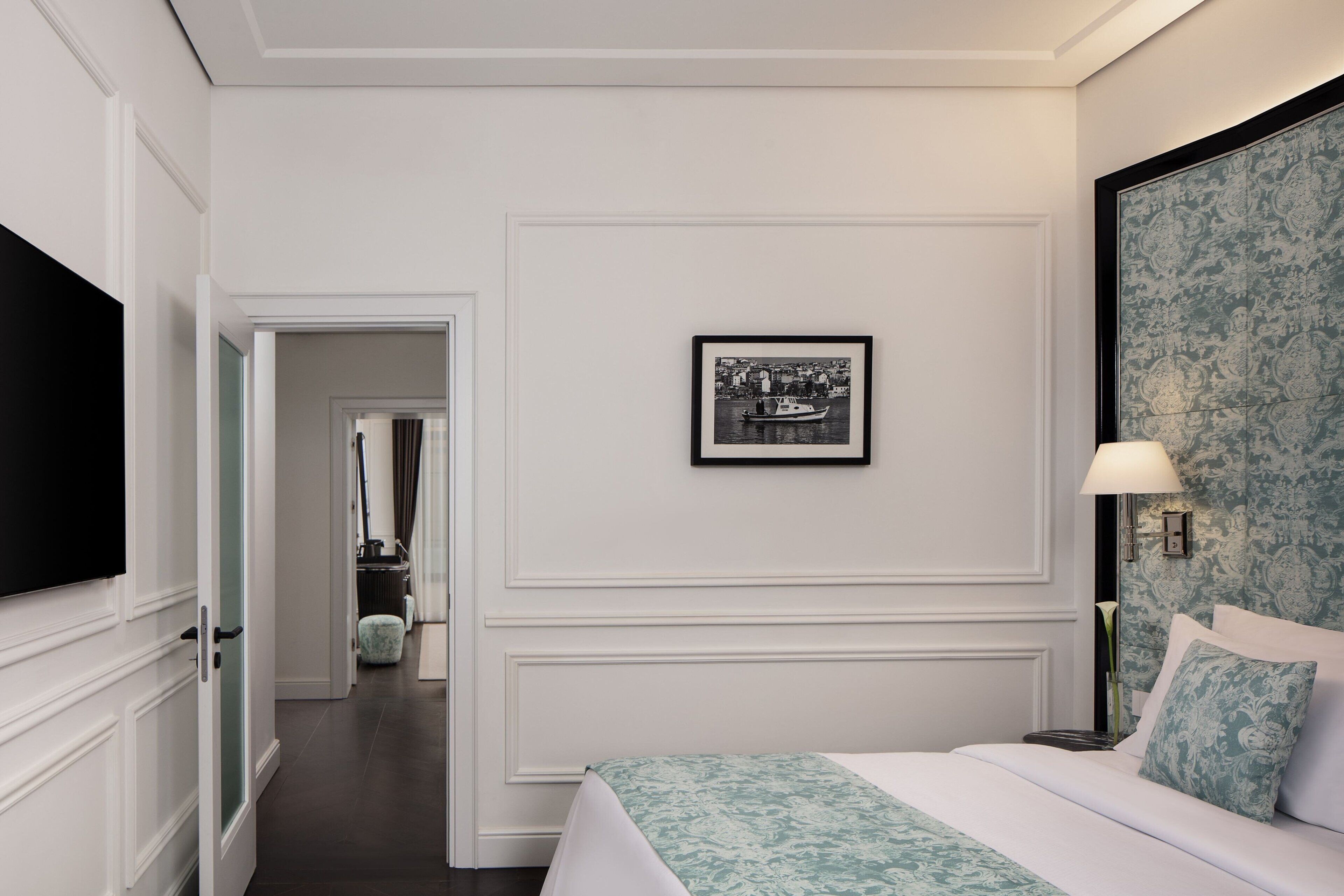 Quarto de hotel com cama, televisão, quadro na parede e mesa de cabeceira com abajur.