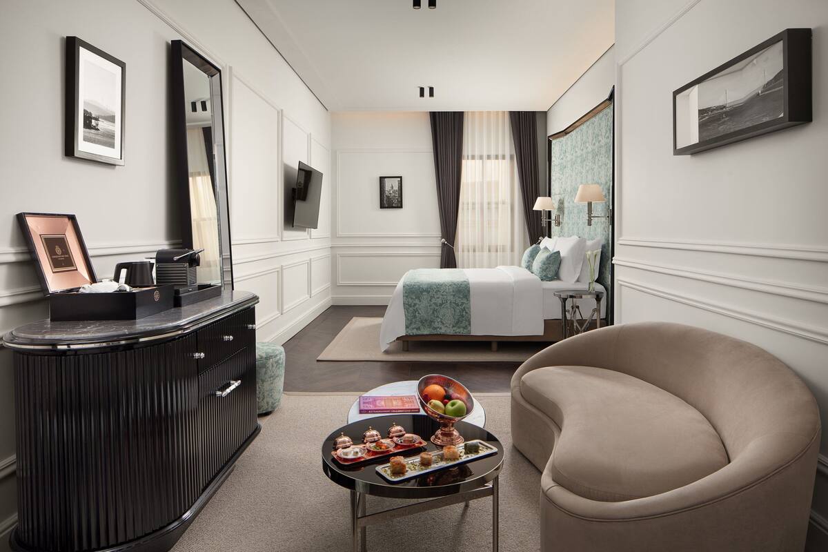 Habitación de hotel con una cama, zona de estar con una silla, una mesita con aperitivos, cómoda y televisor.