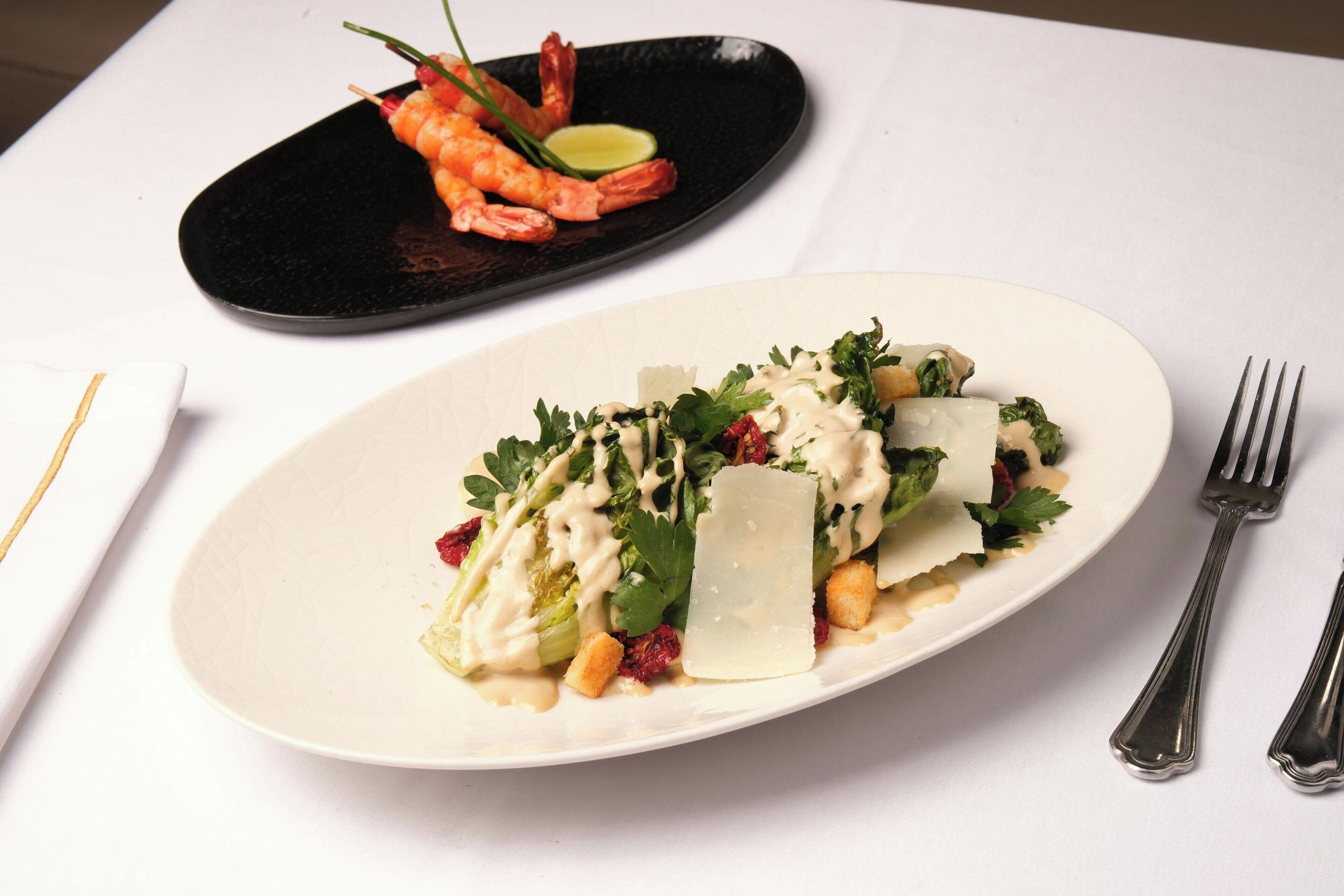 Een wit bord met een salade, een zwart bord met garnalen en een vork op een witte tafel.