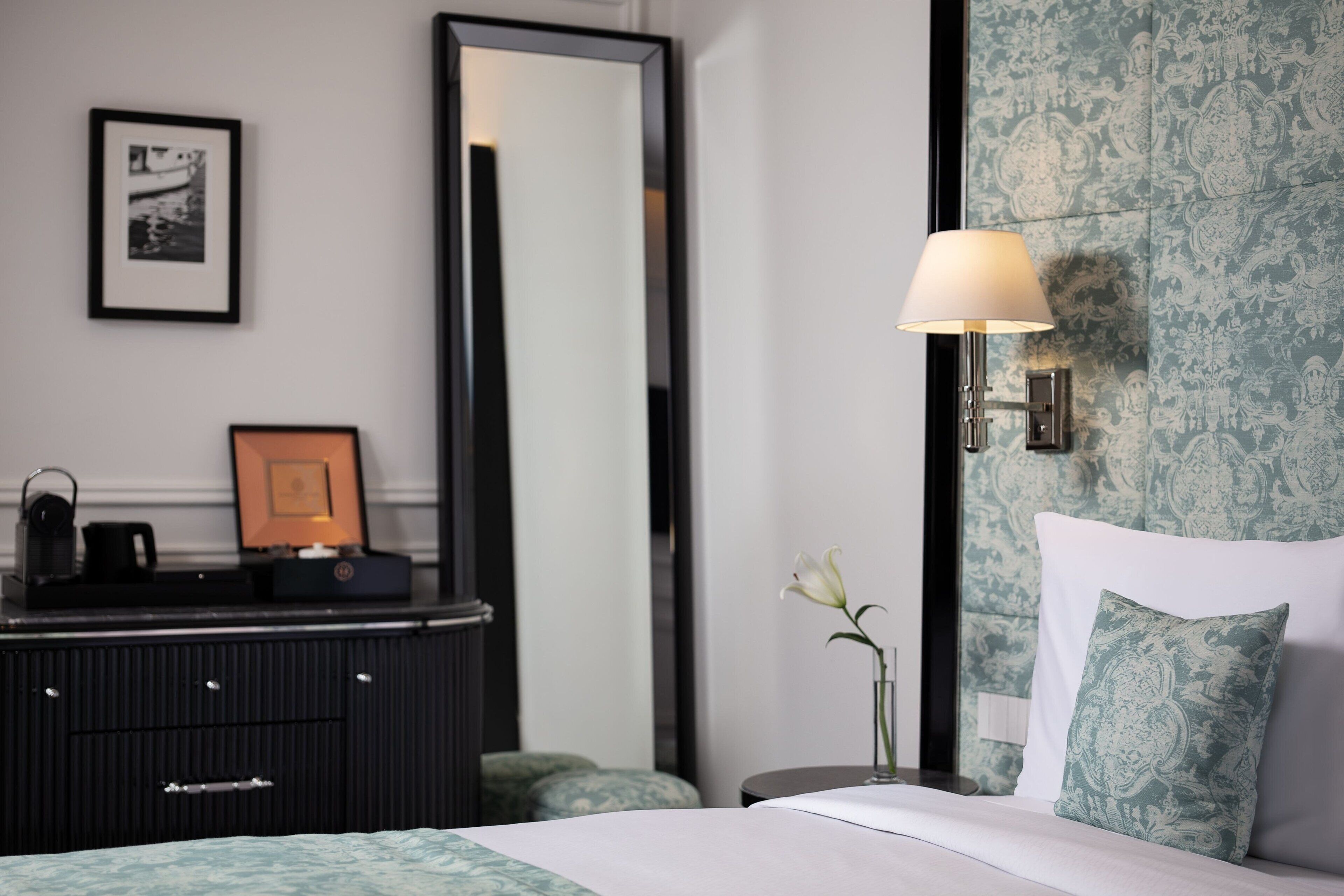 Une chambre d’hôtel avec un lit, une commode, un miroir, une lampe fixée au mur et un tableau encadré accroché au mur.