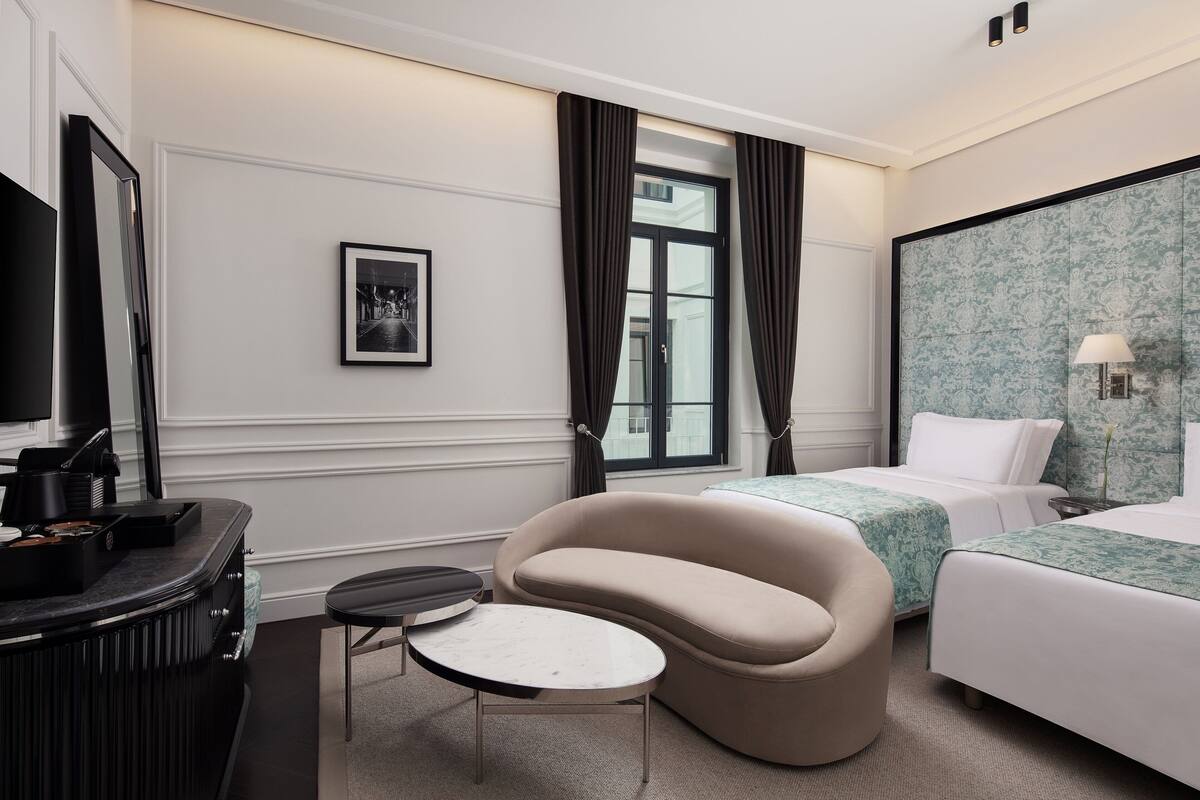Habitación de hotel con un sofá curvo, dos camas individuales, una mesita redonda, un televisor y un cuadro en la pared.