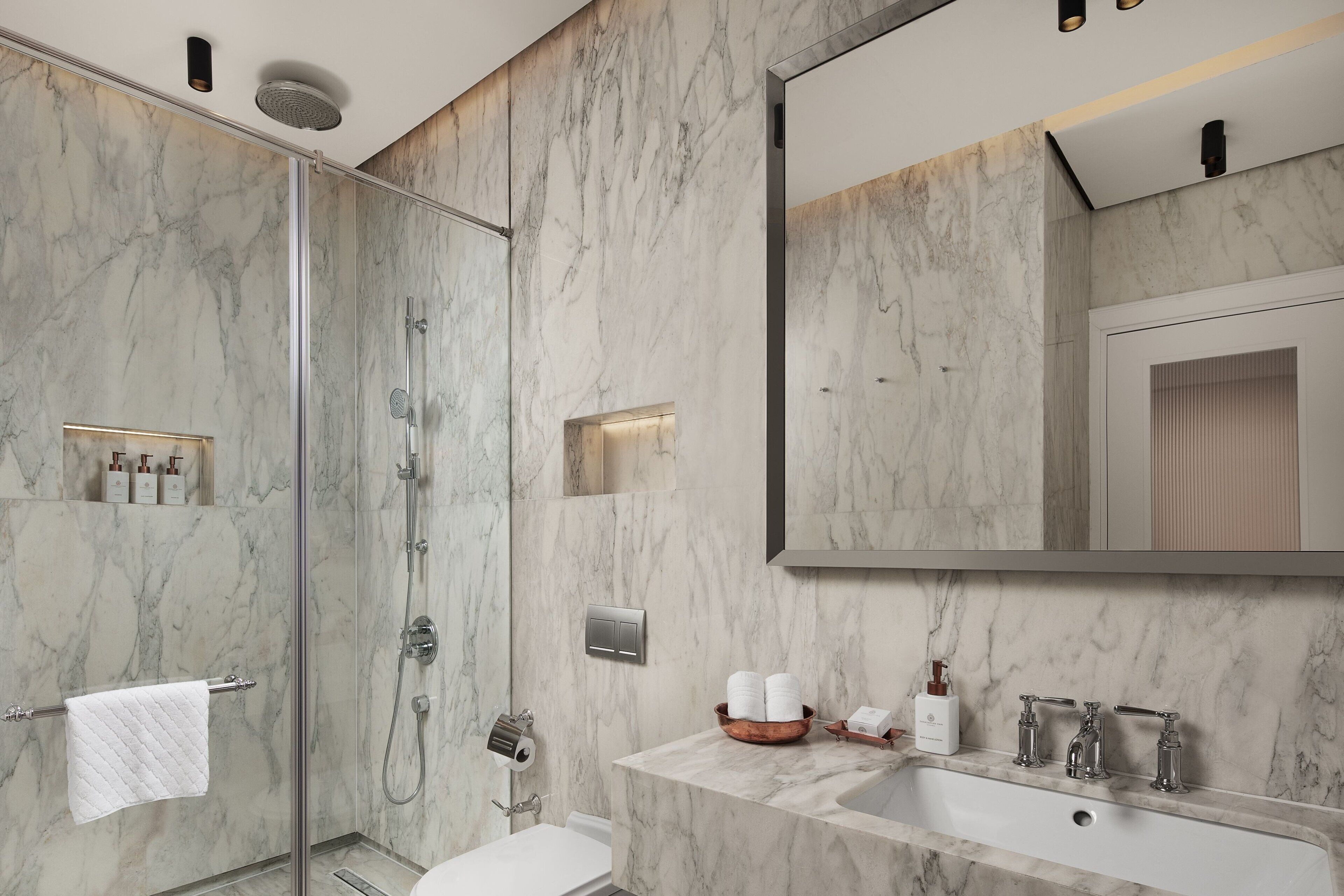 Une salle de bain moderne dotée d’une douche à parois de verre, d’un mur en marbre, d’un grand miroir et d’un lavabo blanc avec deux robinets.