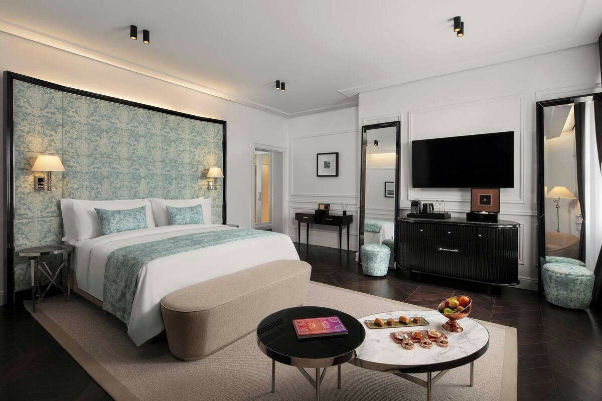 Una habitación de hotel moderna con una cama grande, un televisor de pantalla plana, una mesa de comedor con fruta y una pequeña mesita auxiliar.