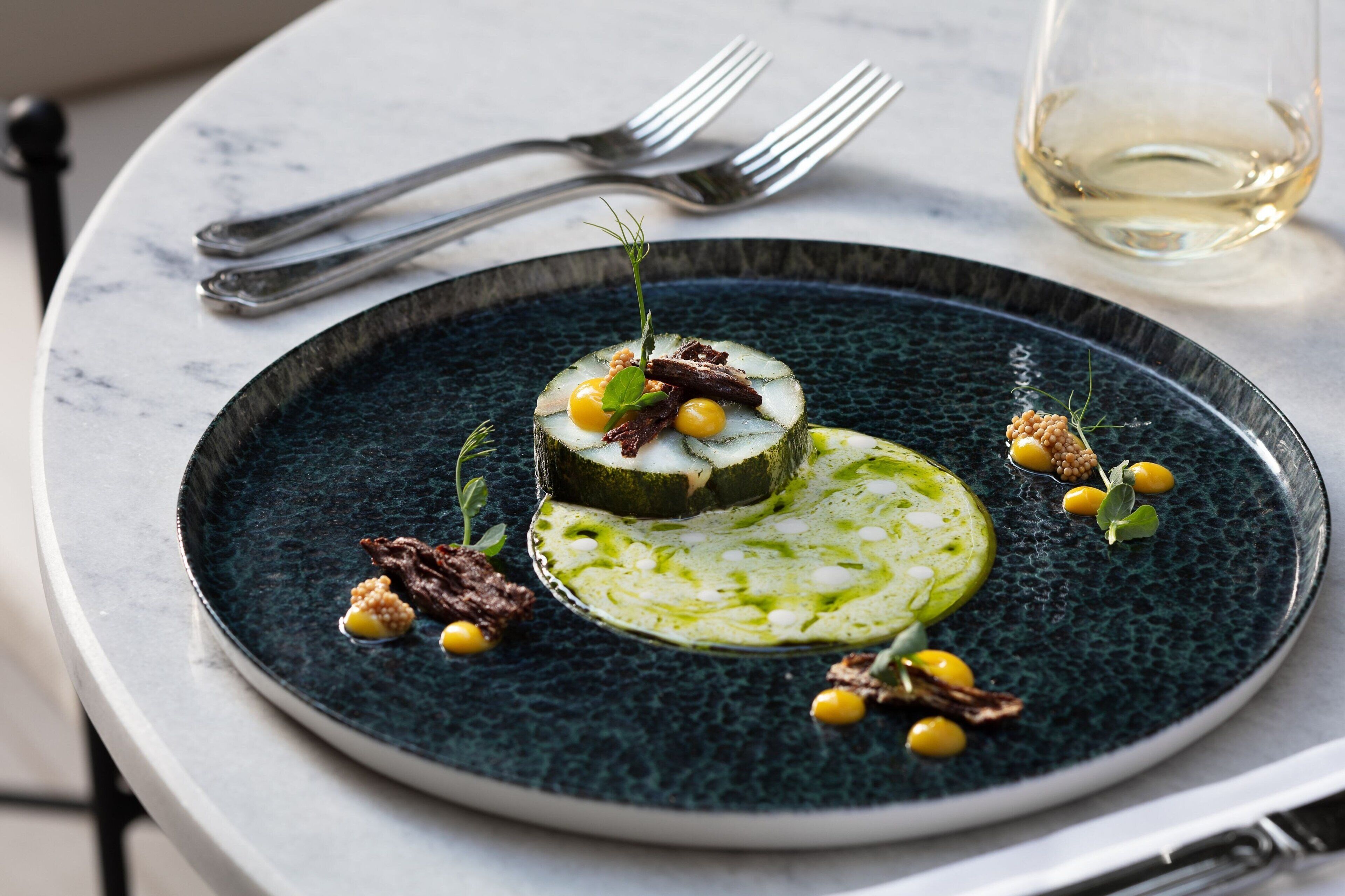 Une assiette de nourriture sur une assiette bleu foncé, un verre de vin blanc et deux fourchettes sur une table en marbre.