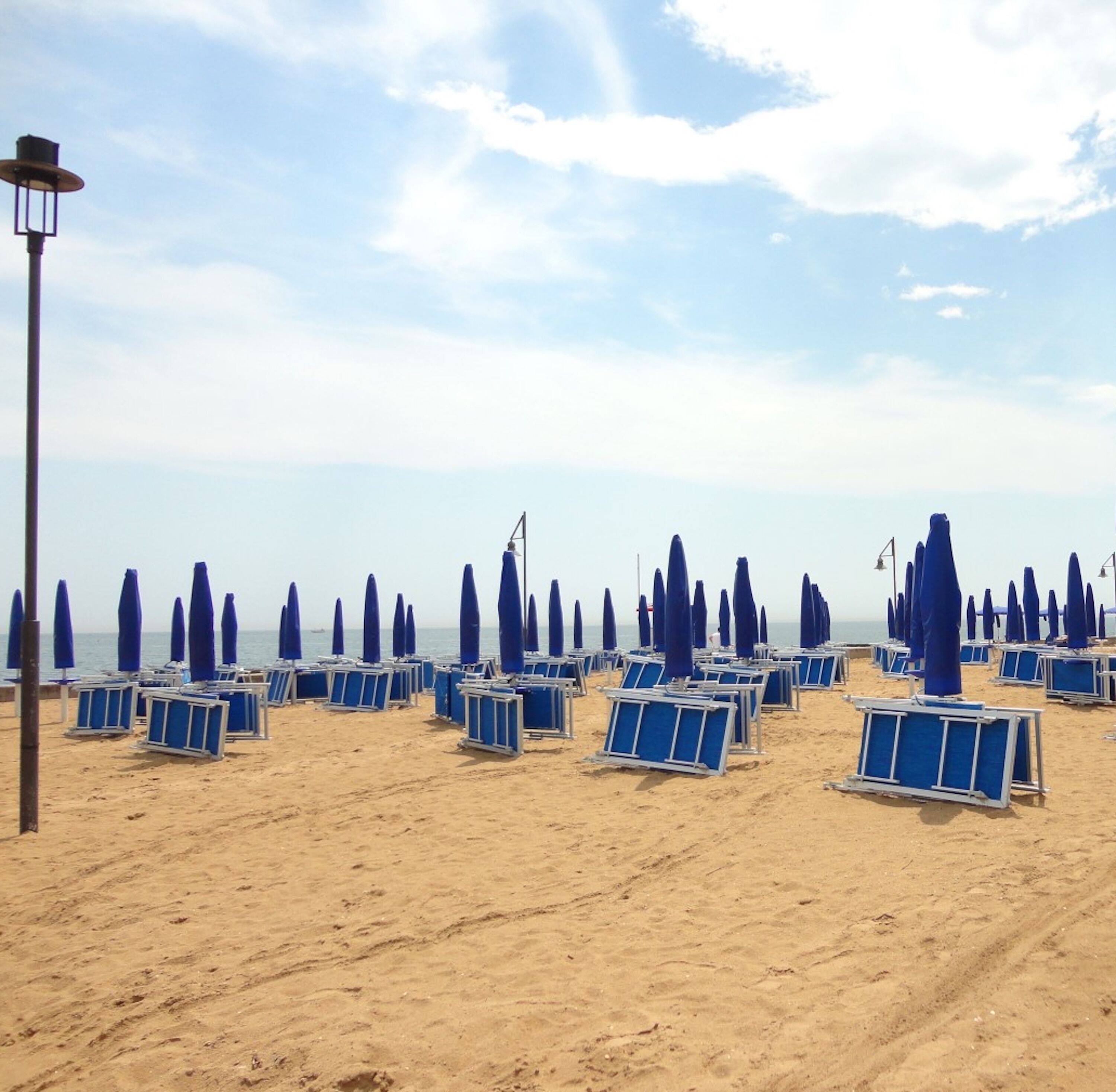 A beach with rows of blue beach chairs and umbrellas, a lamp post, and a clear sky.