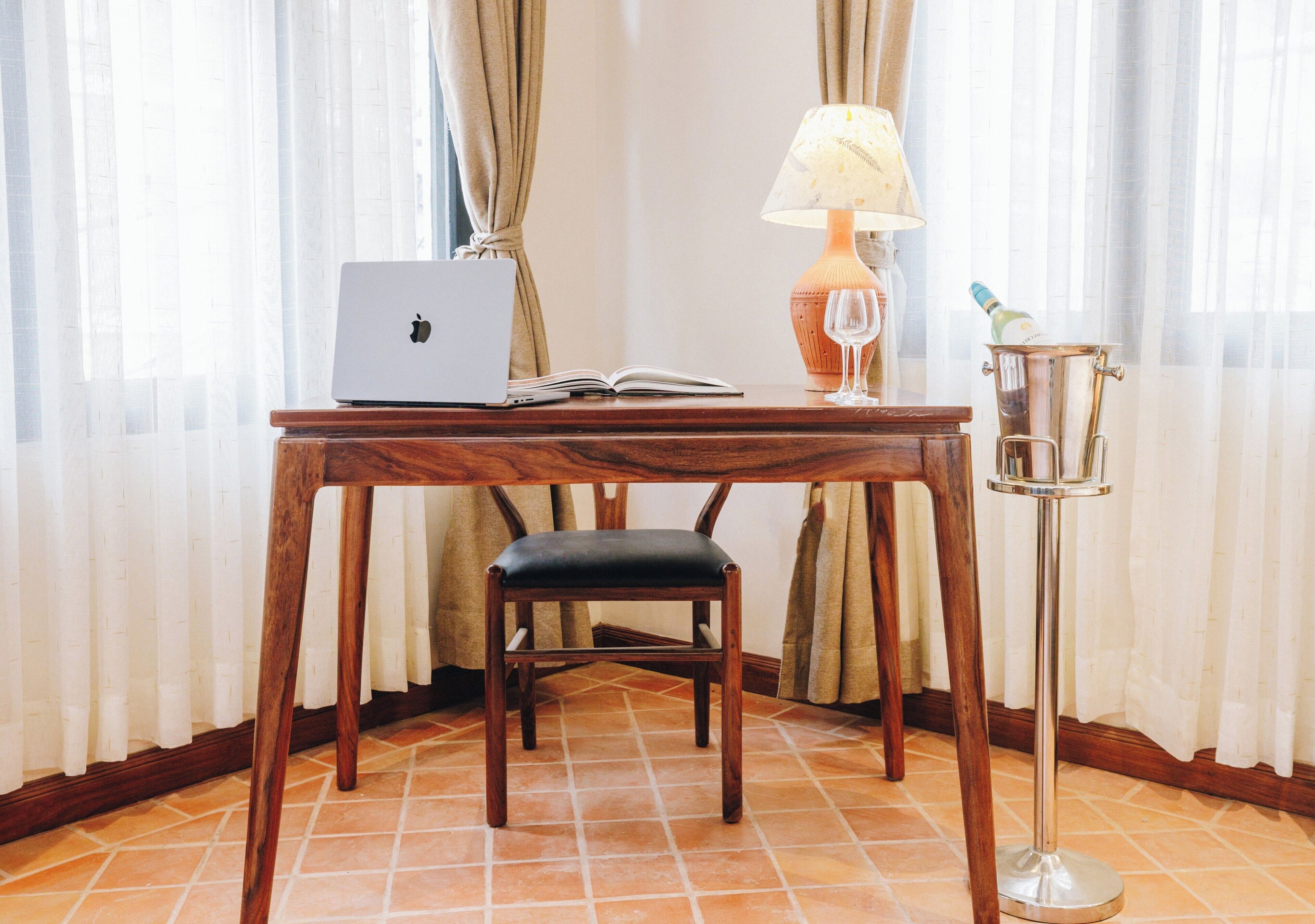 A wooden desk with a laptop, an open book, a wine glass, and a champagne bottle on a table.