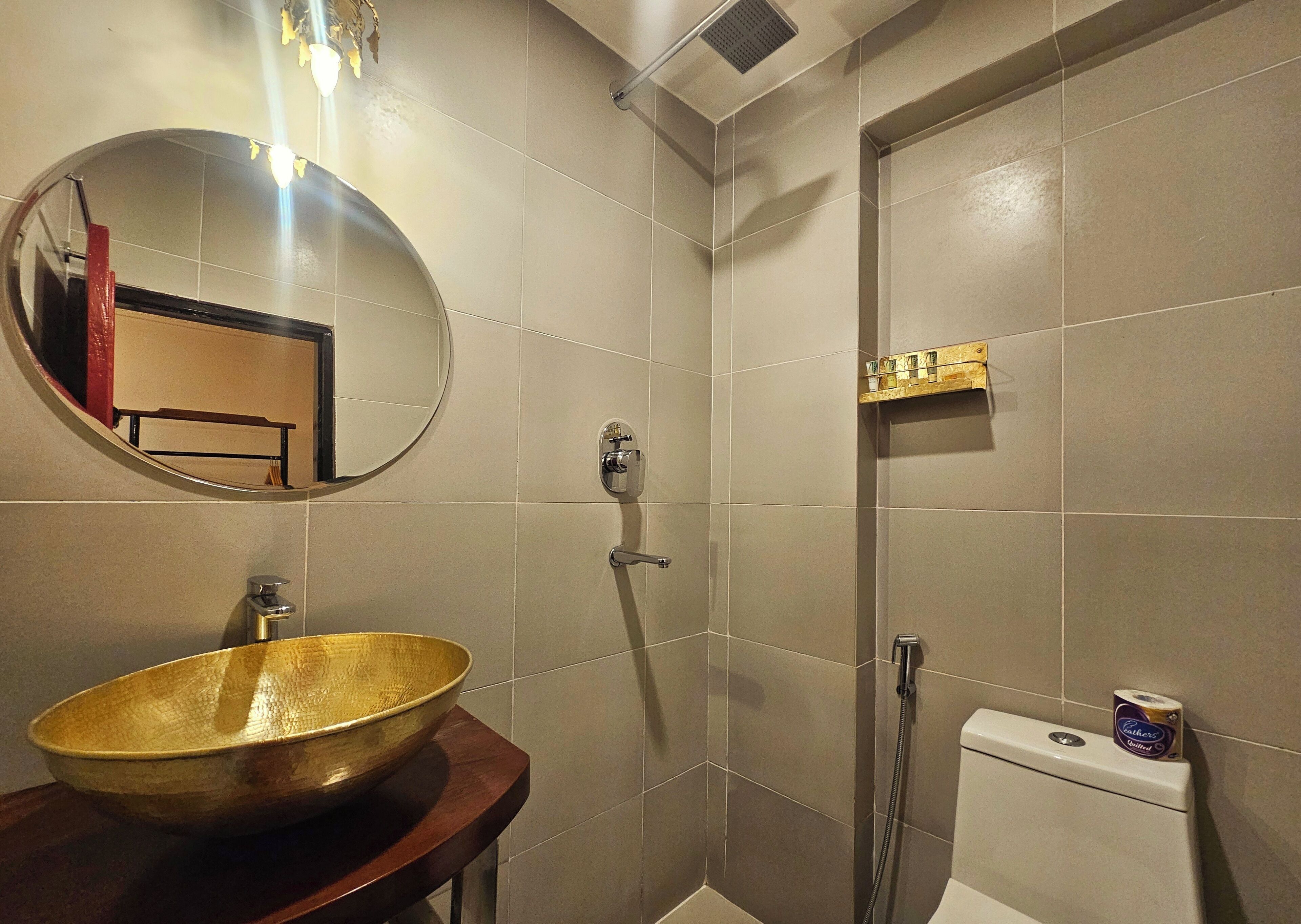 Ein Badezimmer mit rundem Spiegel, einem großzügigen goldfarbenen Waschtisch und einer Dusche sowie Toilette mit Toilettenpapierhalter.