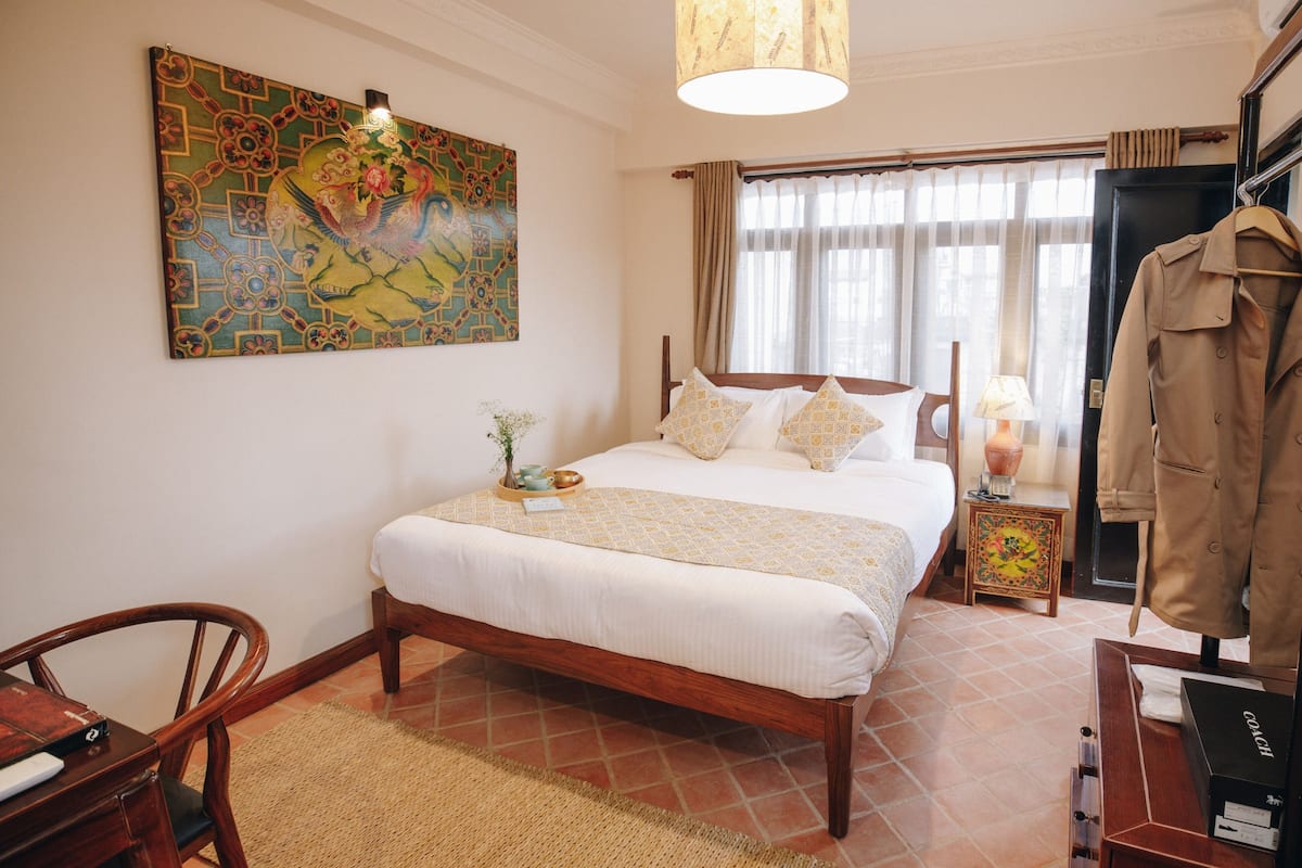 Un dormitorio ordenado con una cama grande, una silla de madera, una mesita con una lámpara y un perchero.