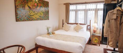 A neatly arranged bedroom with a large bed, a wooden chair, a small table with a lamp, and a coat rack.