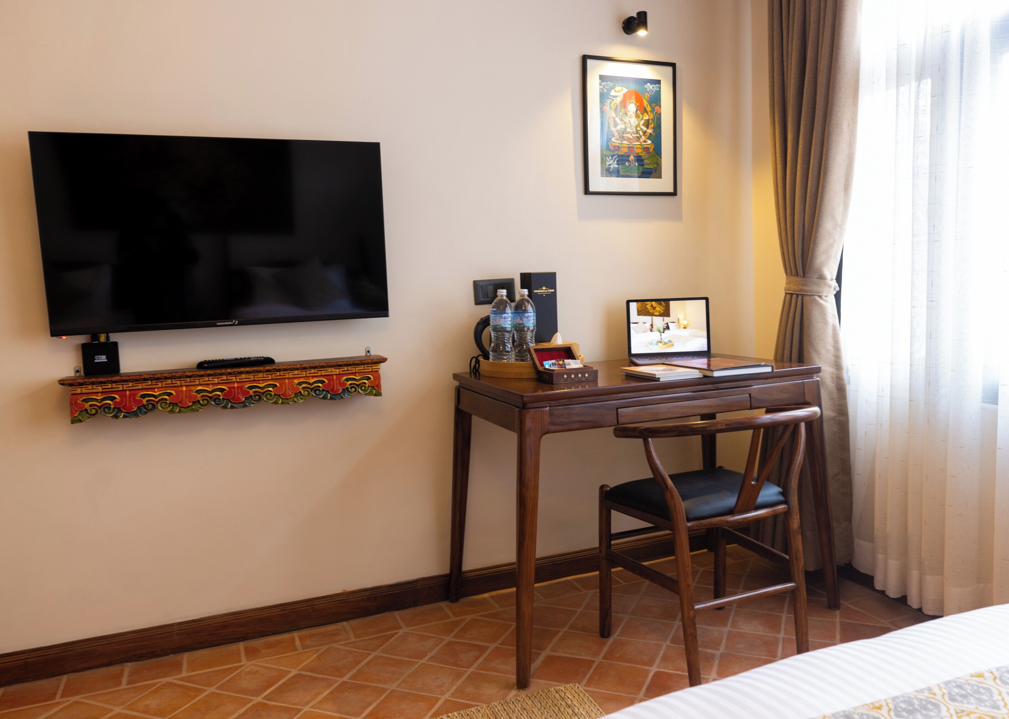 A hotel room with a flat-screen TV, a desk with a laptop, a chair, a bed, a decorative shelf, and a framed picture on the wall.