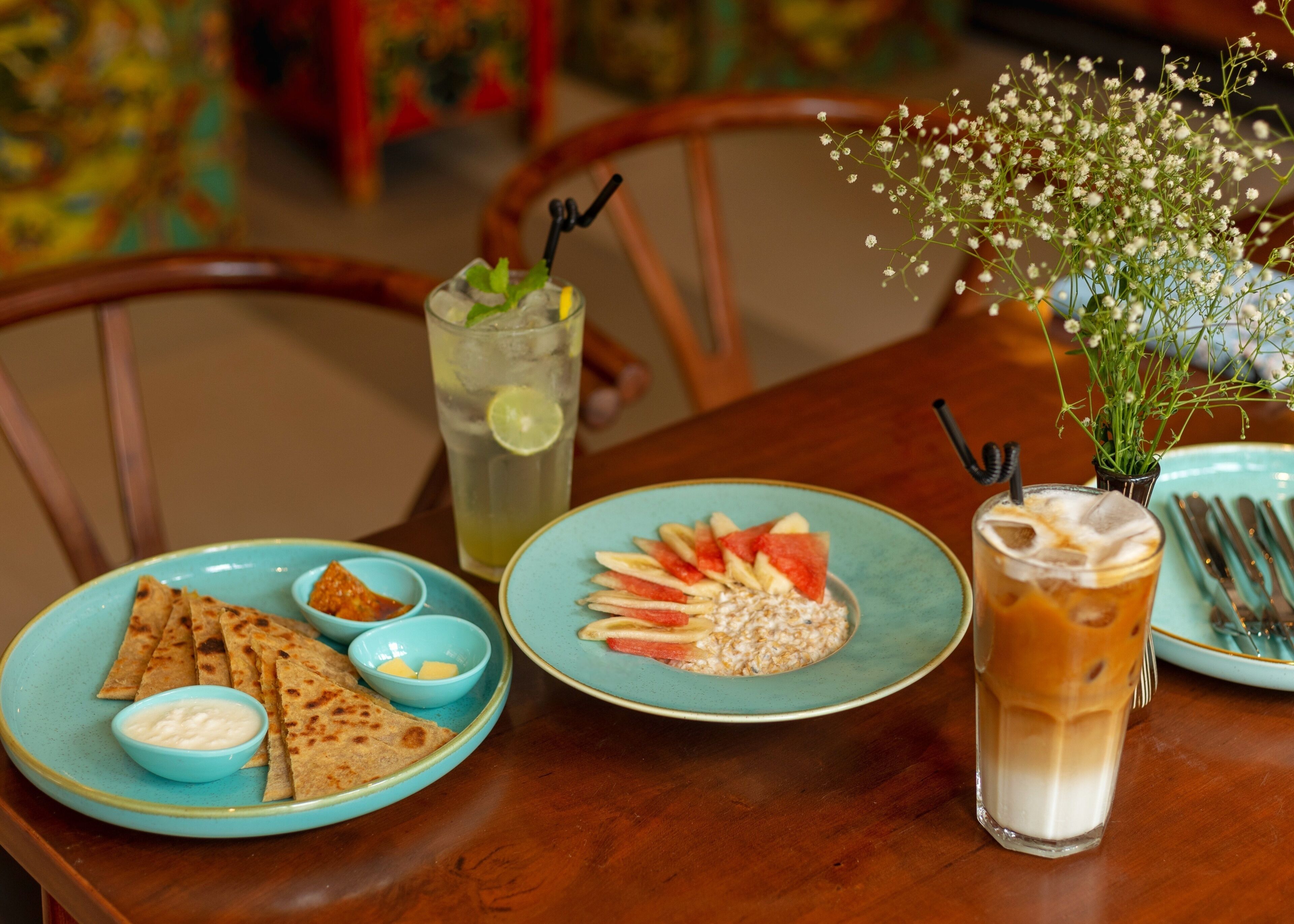 A table with a plate of flatbreads, a bowl of yogurt, a plate with sliced fruits and oats, a glass of iced drink, and a glass of iced coffee.