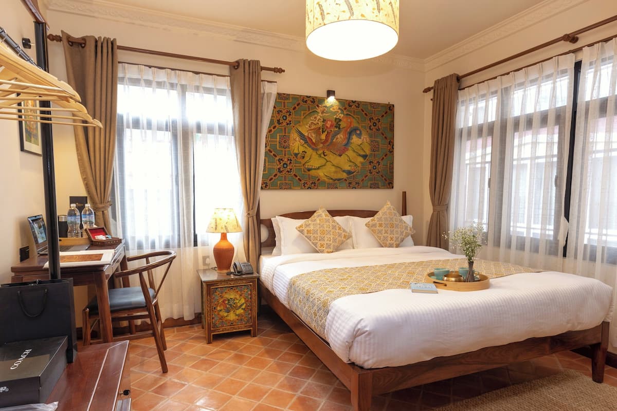 Habitación de hotel con cama, escritorio con silla, lámpara, un cuadro en la pared y cortinas.