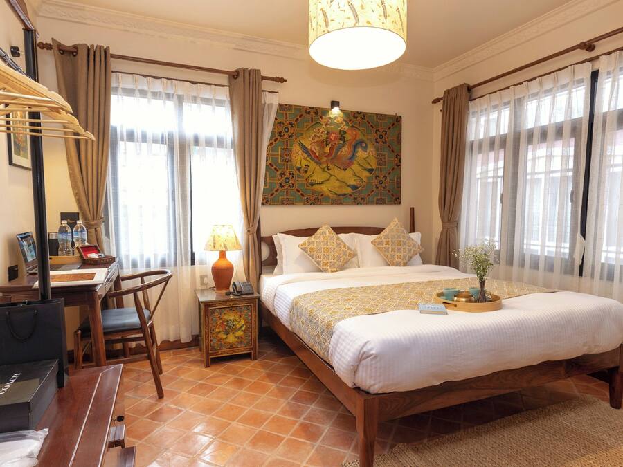 Habitación de hotel con cama, escritorio con silla, lámpara, un cuadro en la pared y cortinas.