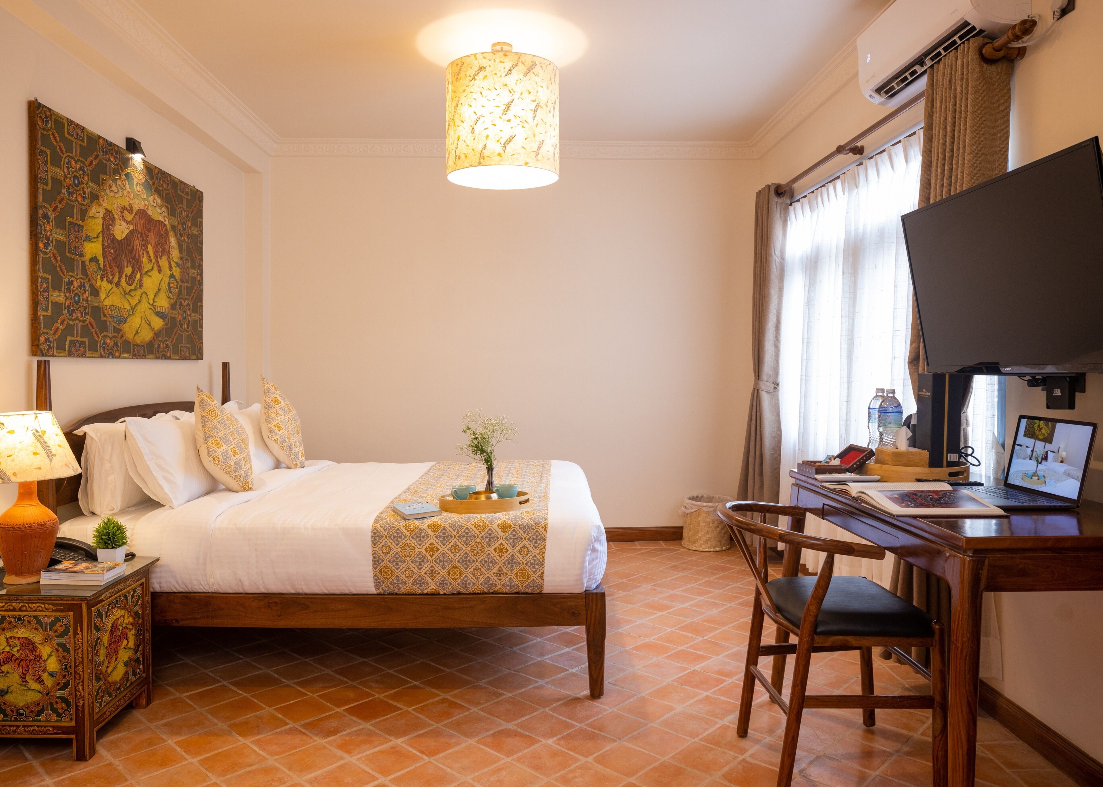 A hotel room with a bed, a desk with a laptop, a lamp, and a painting on the wall.
