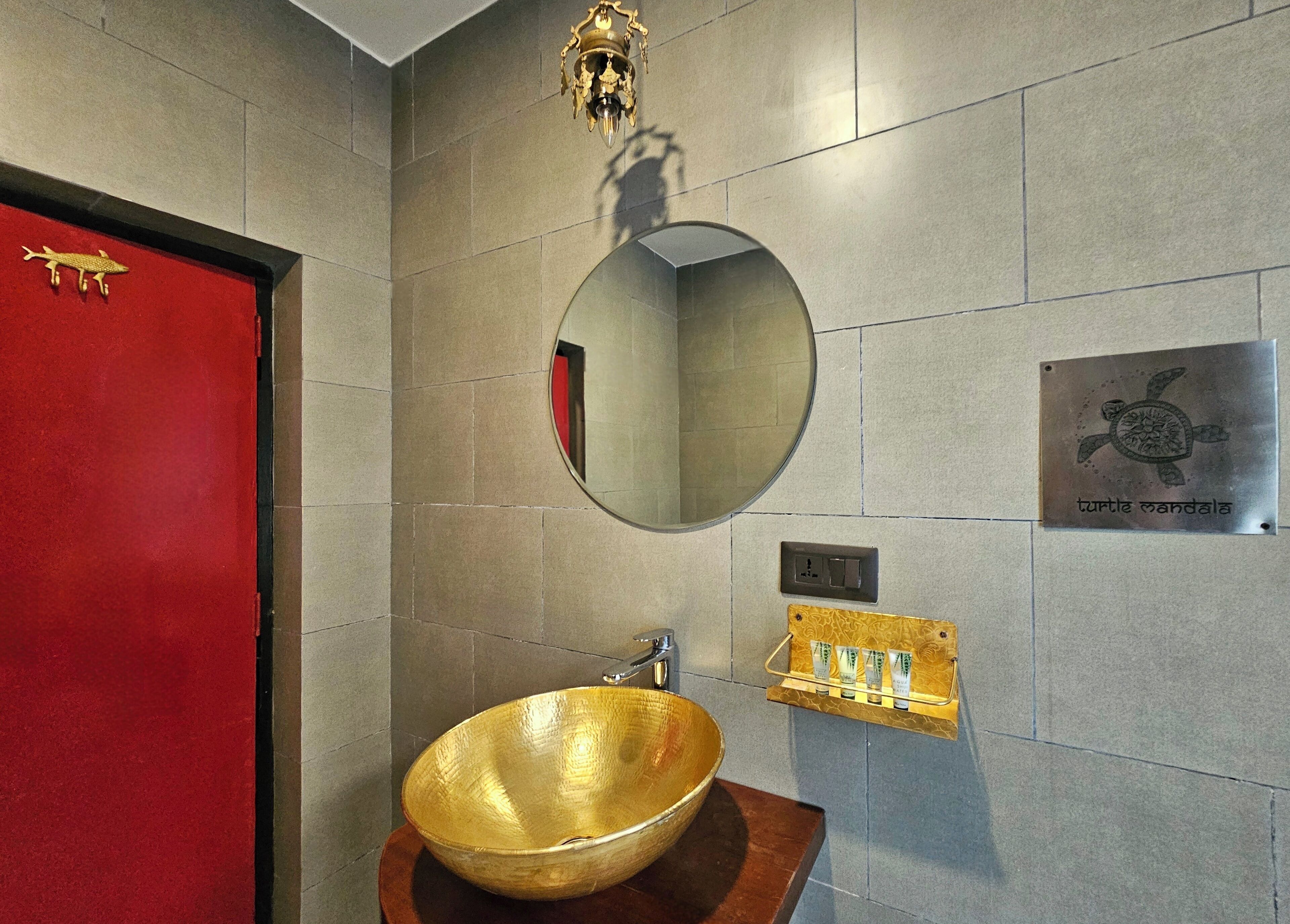 A bathroom with a round mirror, a gold bowl sink, a red door, and a plaque with a turtle design.