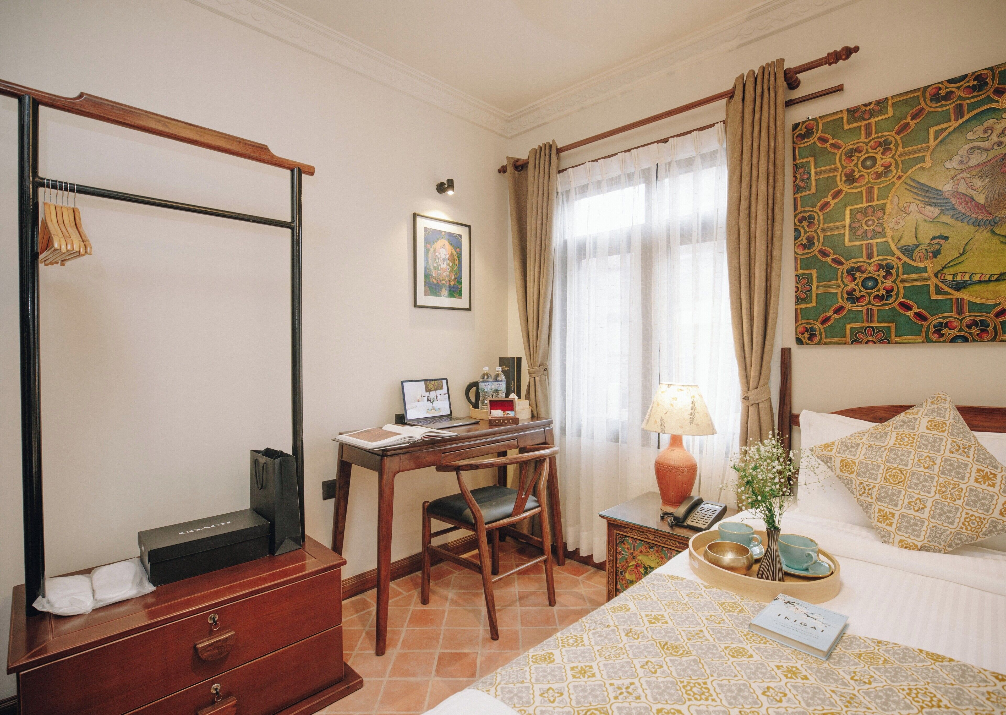 A hotel room with a wooden desk, a laptop, a chair, a bed with patterned pillows, a bedside table with a lamp, and a painting on the wall.