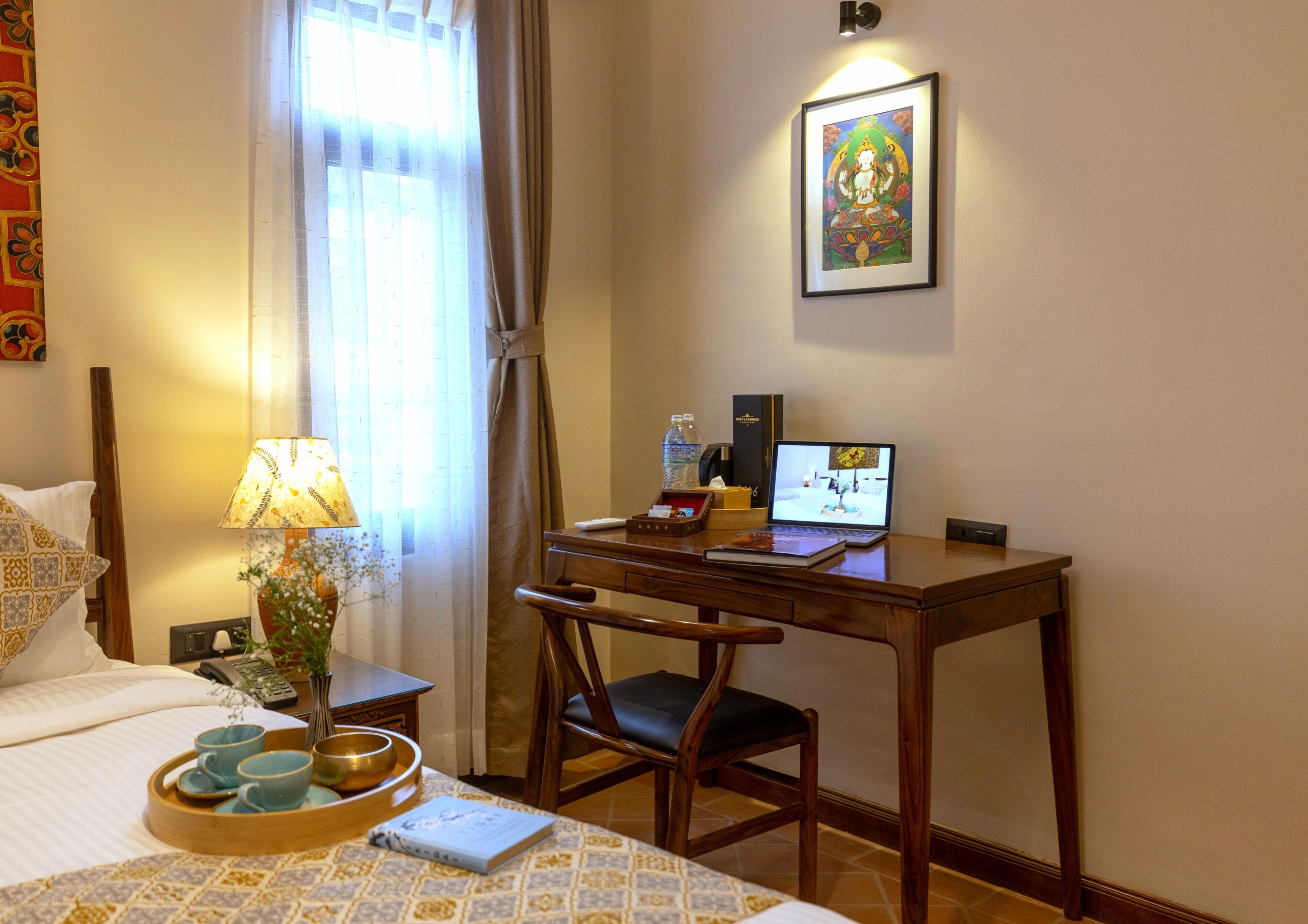 A hotel room with a bed, a desk with a laptop, a chair, a bedside table with a lamp, a tray with cups and a bowl, and a framed picture on the wall.