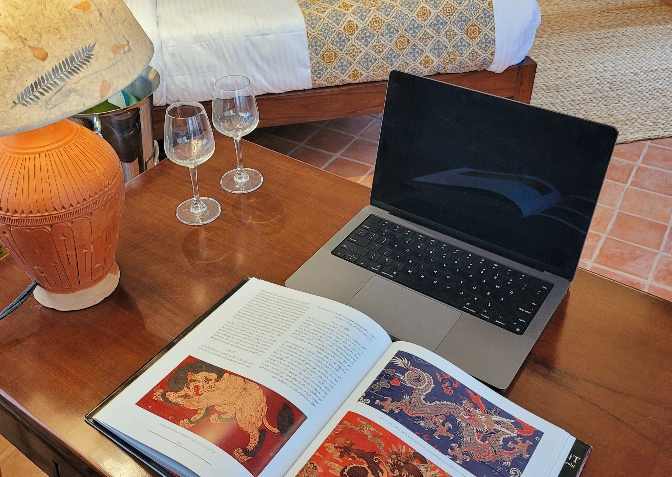 A laptop on a wooden table with two open books, a lamp, and three wine glasses.