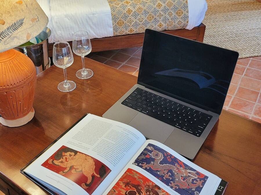 A laptop on a wooden table with two open books, a lamp, and three wine glasses.
