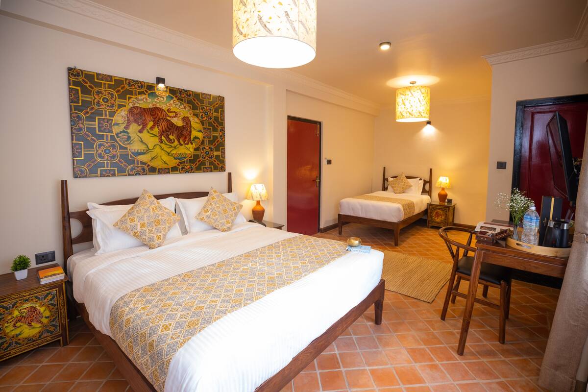 Una habitación de hotel con una cama grande, una cama más pequeña, un escritorio con silla, y una mesa de comedor. Hay un cuadro en la pared y una puerta roja.