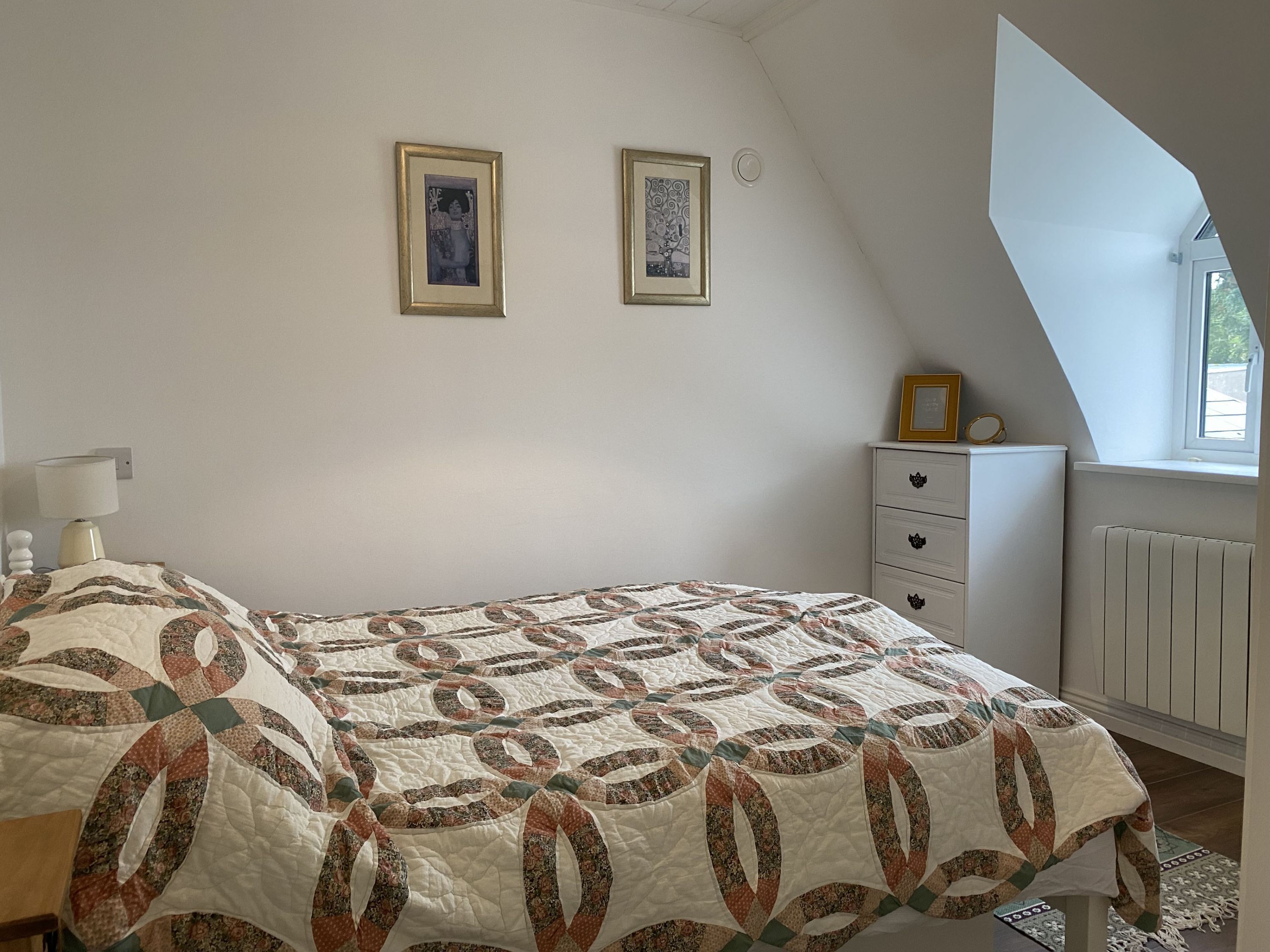 Een slaapkamer met een bed, een dressoir, twee ingelijste foto’s aan de muur en een raam met uitzicht.