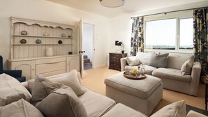 Ein Wohnzimmer mit einem beigefarbenen Sofagarnitur, einem Couchtisch mit Tablett, einem weißen Schrank mit dekorativen Tellern und einem Fenster mit Vorhängen.