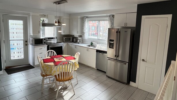 A compact kitchen with white cabinets, a stainless steel refrigerator, a microwave, and a stove. There is a dining table with two chairs and a red placemat.