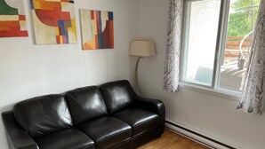 A black leather sofa in a room with wooden flooring, a window with a view of the outdoors, and abstract paintings on the wall.