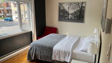 A hotel room with a bed, a bedside table with a lamp, a window with a view of the street, and a painting on the wall.
