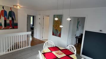 A dining area with a table set for four, white chairs, a red and yellow tablecloth, and a painting of a tree on the wall.