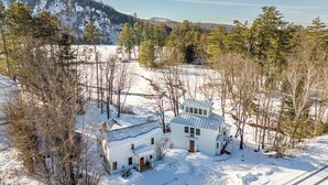 A white house with a red door, surrounded by snow-covered ground and trees, with a mountainous backdrop. - Apres-Ski House: 20 min to Sunday River and Steam Room (Bryant Pond)