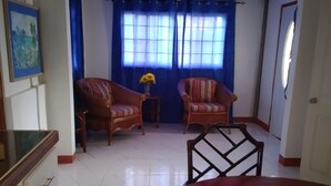 Una sala de estar con una mesa roja, dos sillones de madera, una mesita auxiliar con un jarrón de flores amarillas y un cuadro en la pared.