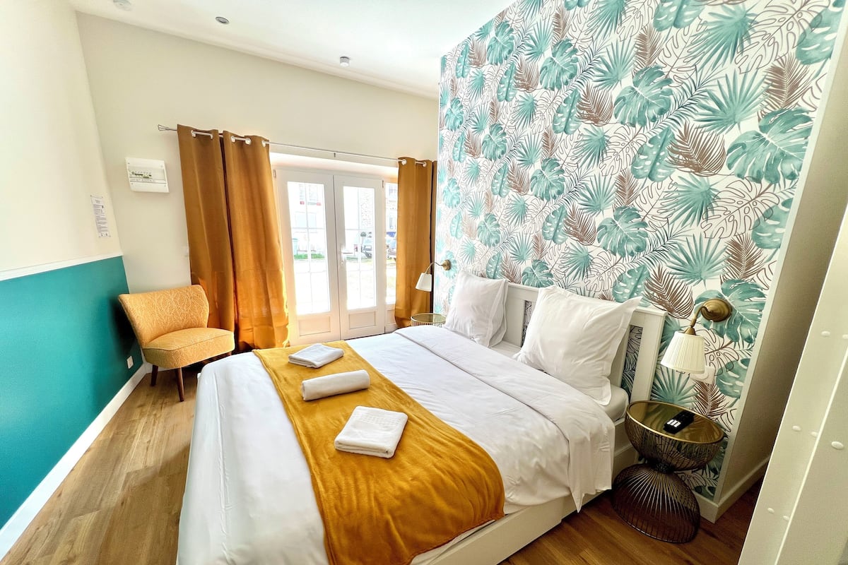 Quarto de hotel com cama, uma cadeira, mesa de cabeceira e janela com cortinas.