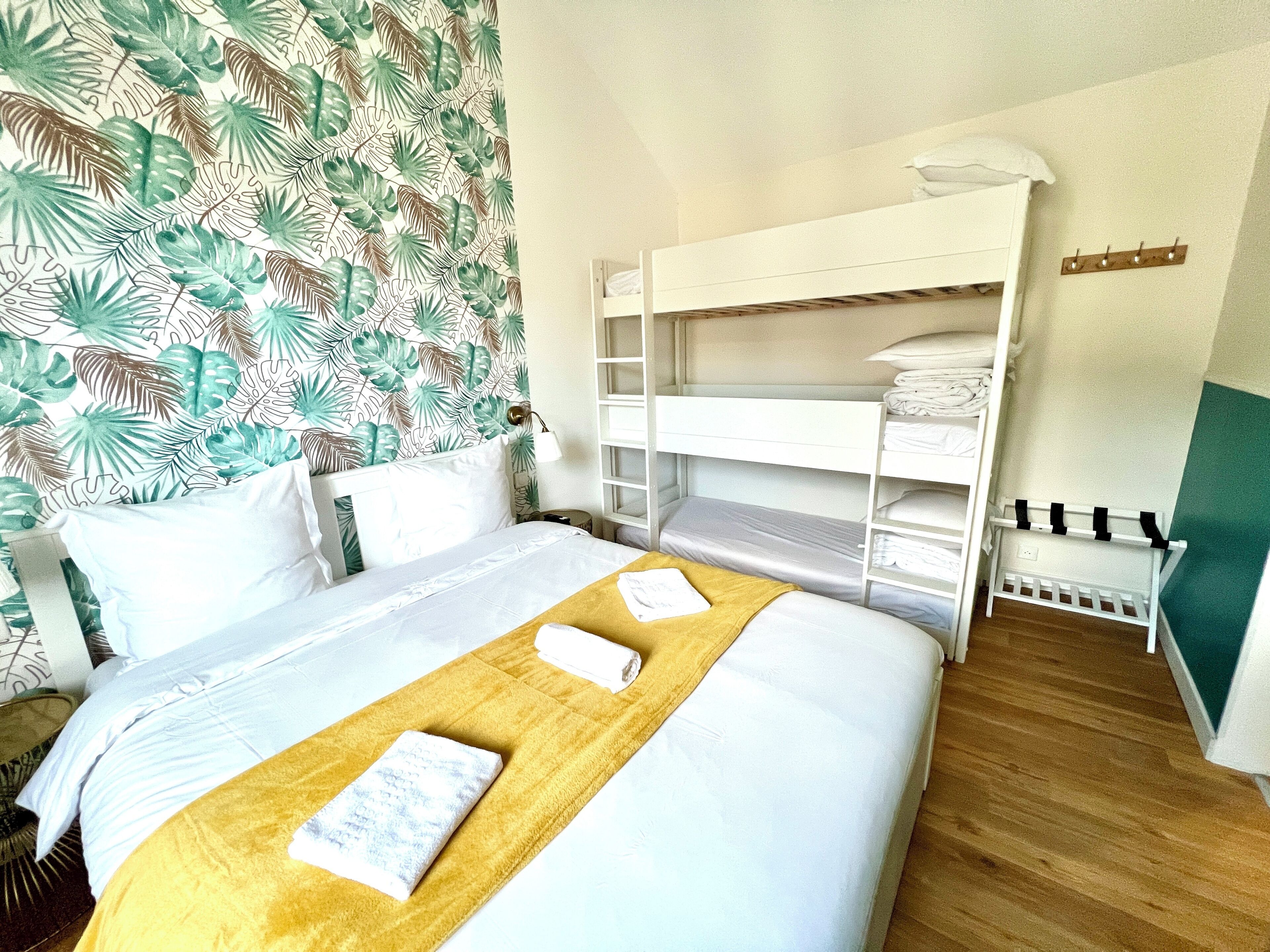 A hotel room with a bunk bed, a wooden bedside table, a bed with a yellow throw, and a tropical leaf-patterned wallpaper.