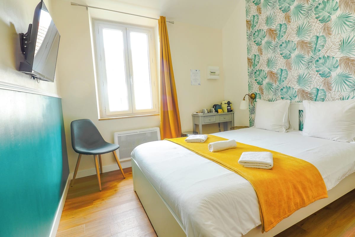 Quarto de hotel com cama, escrivaninha, cadeira, radiador e janela com cortinas.