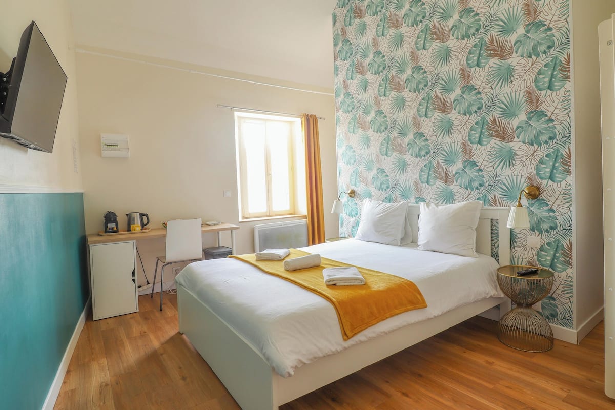 Quarto de hotel com cama, escrivaninha, cadeira, janela com cortinas e parede com padrão de folhas.