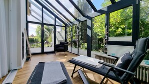 Ein sonnendurchfluteter Raum mit einer großen Glaswand, einer Bank, einem Sessel und Blick auf einen Garten.