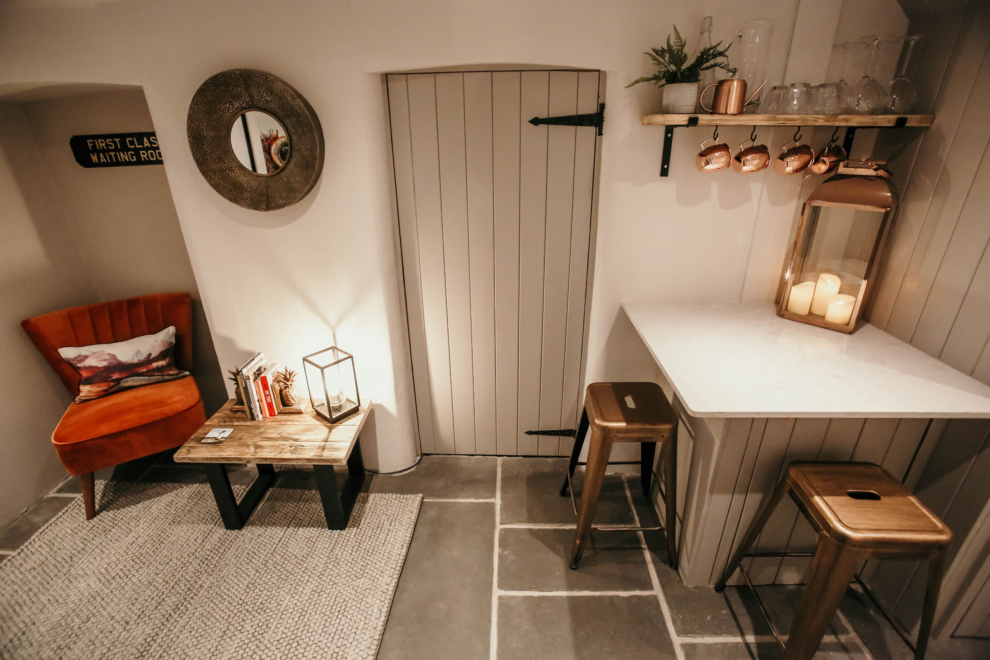 A cozy room with a wooden door, a red chair, a small table with books and a lantern, and a shelf with glassware and copper mugs.