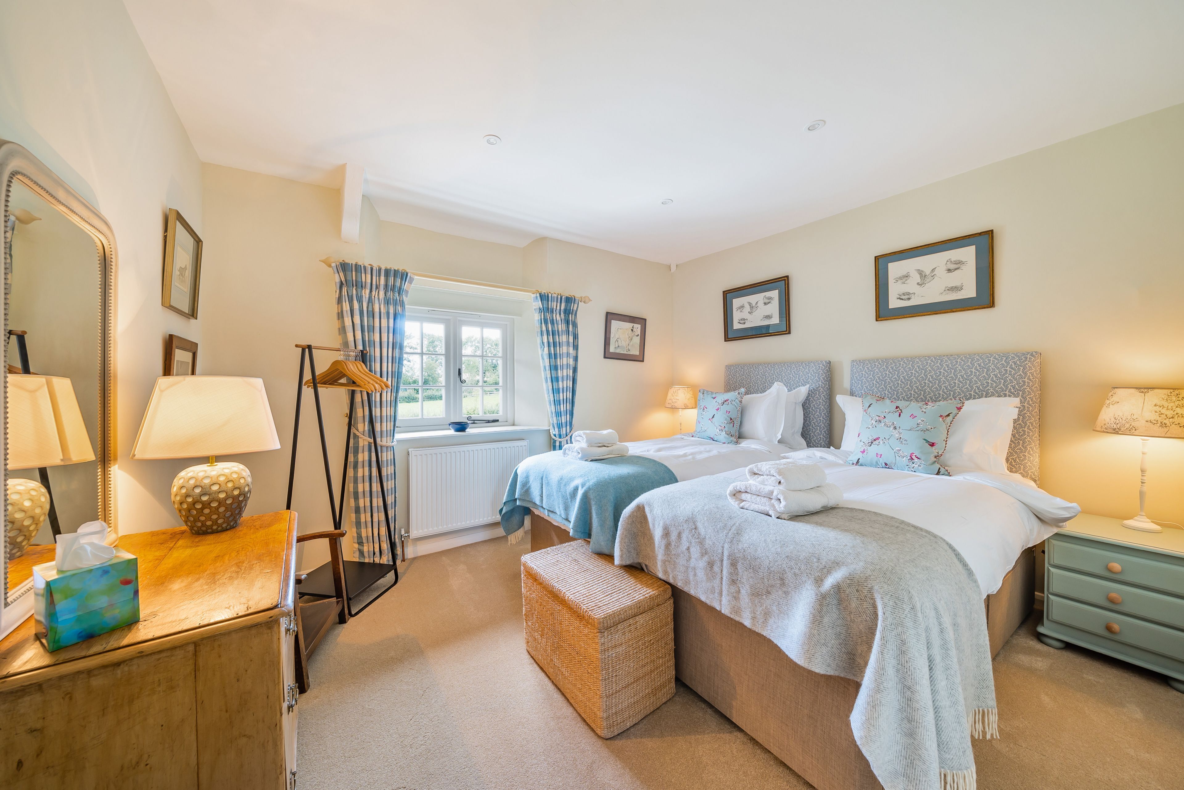 A well-lit bedroom with two beds, a dresser, a mirror, and a window with curtains.