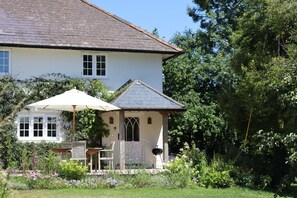 A white house with a tiled roof, a covered porch with a table and chairs, and a garden with various plants and flowers. - Cosy Cottage - Chebbard, Dorchester (Dorchester)