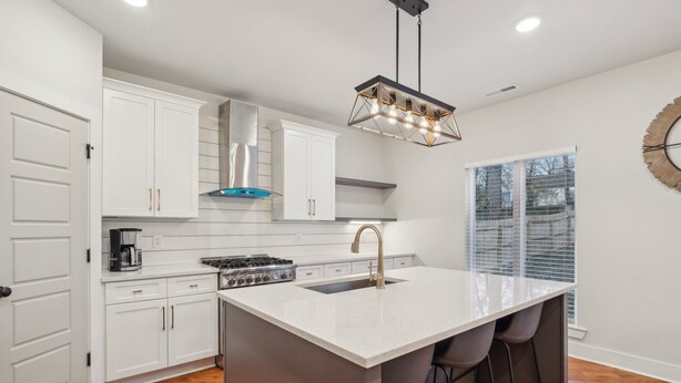 Una cocina moderna con armarios blancos, una isla central con taburetes de bar, una campana extractora de acero inoxidable y una lámpara colgante.