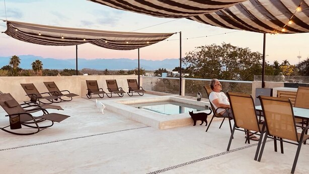 A rooftop pool area with lounge chairs and a person sitting under a striped canopy.