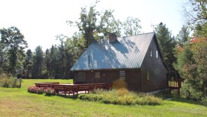Une cabane en bois avec un toit en métal, entourée d’une zone herbeuse où se trouvent des bancs et une table de pique-nique.