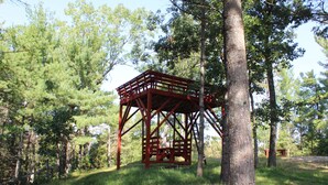 Un gazebo en bois rouge, situé dans une zone boisée, entouré de grands arbres et d’une pelouse verdoyante.