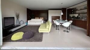 A modern living room with a sofa, a dining table with chairs, a TV, and a kitchen area. - Herdade dos Salgados, T1 6B_1C, Vila das Lagoas (Albufeira)