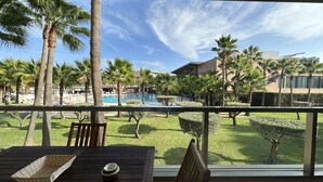 A poolside view with palm trees, a swimming pool, and a building in the background. - Herdade dos Salgados, T1 6B_1C, Vila das Lagoas (Albufeira)