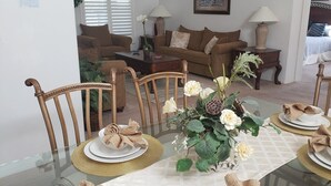 Uma área de jantar com uma mesa de vidro posta para quatro pessoas, rodeada por cadeiras de madeira. Um vaso com flores e um objeto decorativo estão sobre a mesa.
