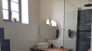 A modern bathroom with a round mirror, a white sink, a wooden stool, and a glass-enclosed shower area.