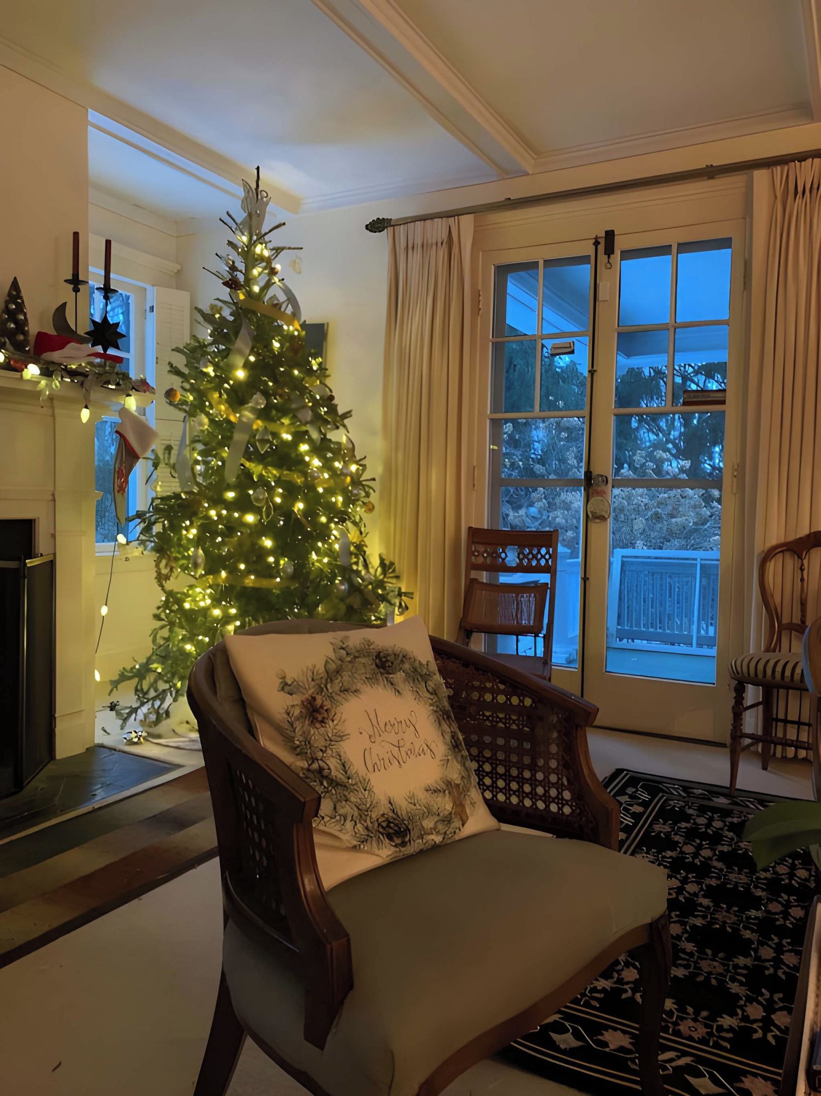 A living room with a Christmas tree, a fireplace, a large window with curtains, and a wooden chair with a cushion.